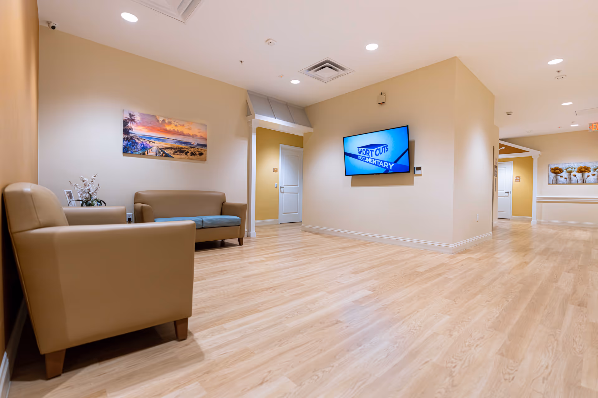 Bright senior living facility common seating area with beige sofas, wall-mounted TV, artwork, and wood flooring.