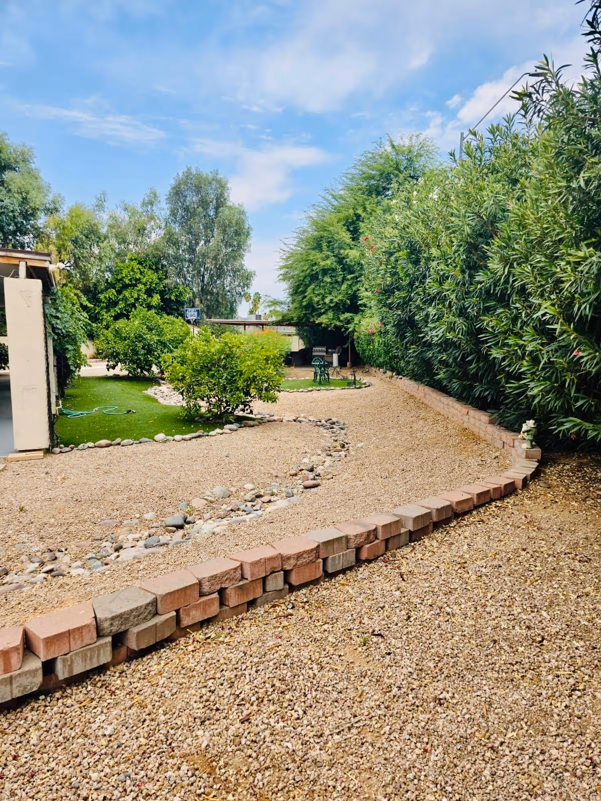 A landscaped outdoor garden area with a gravel pathway bordered by bricks and rocks, green bushes, trees, and a small seating area with a table and chairs under a partly cloudy sky.