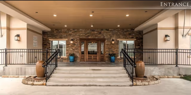 Entrance to a building with stone wall facade, wooden double doors, two windows, black metal railings on either side of concrete steps, large ceramic pots with plants, and wall-mounted lantern lights under a covered porch.