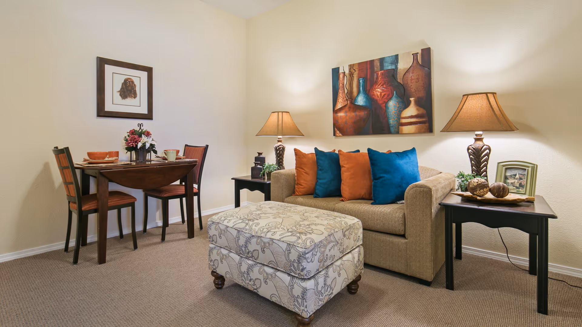 A cozy living room area with a beige sofa adorned with blue and orange throw pillows, two dark wooden side tables each with a lamp, a patterned ottoman in front of the sofa, and a small dining table with two chairs set against a light-colored wall. The walls are decorated with framed artwork, including a painting of vases above the sofa and a portrait of a dog above the dining table.