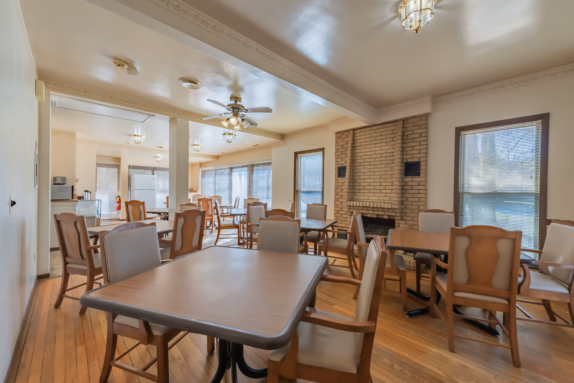 A spacious dining room with multiple wooden tables and cushioned chairs arranged neatly. The room features large windows with blinds, a brick fireplace, ceiling fans with lights, and hardwood flooring. The background shows a kitchen area with a refrigerator and other appliances.