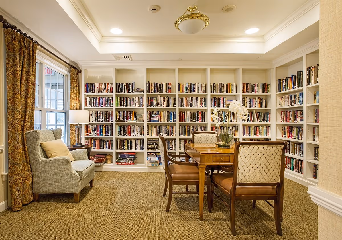A cozy library room with white built-in bookshelves filled with books along the back and right walls. In the center, there is a wooden table with four upholstered chairs around it. To the left, a comfortable armchair with a beige cushion sits next to a window with patterned curtains and a table lamp. The room has a beige carpet and a ceiling light fixture.