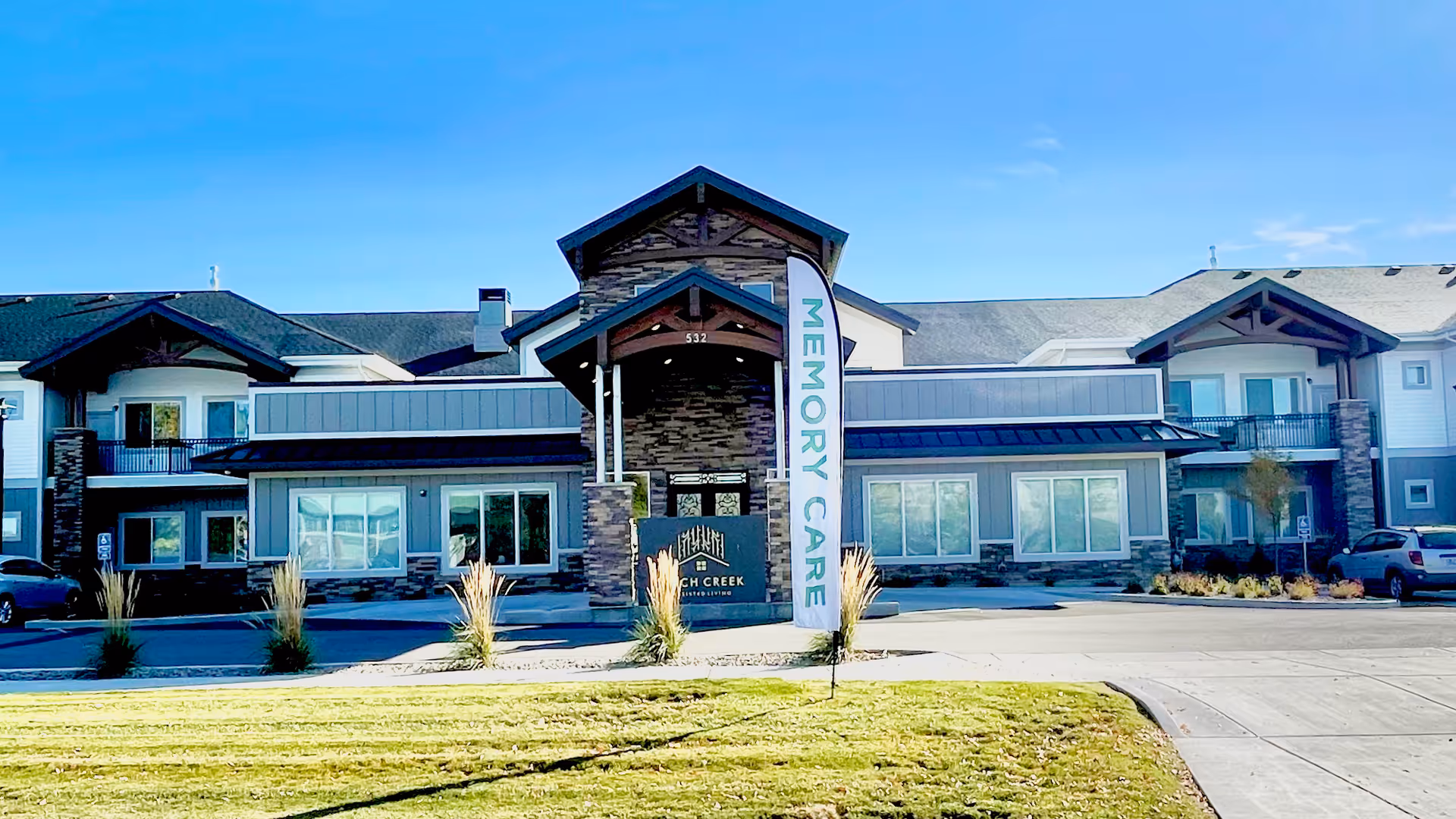 Front exterior view of Birch Creek Assisted Living of Smithfield building under a clear blue sky, featuring a central entrance with a peaked roof, stone and wood accents, multiple windows, and a flag sign that reads 'MEMORY CARE'. Cars are parked on either side of the entrance.