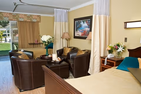 A bright senior living bedroom with a bed, bedside table and flowers, a seating area of leather chairs around a coffee table, and sliding glass doors to an outdoor area.