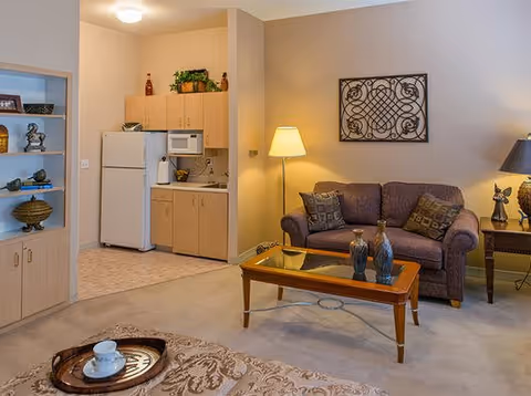 A cozy living area with a small kitchen in the background. The living room features a purple loveseat with decorative pillows, a wooden coffee table with vases, a side table with a lamp and figurine, and a wall decoration. The kitchen area includes a refrigerator, microwave, sink, and cabinets. A built-in shelving unit with decorative items is visible on the left.