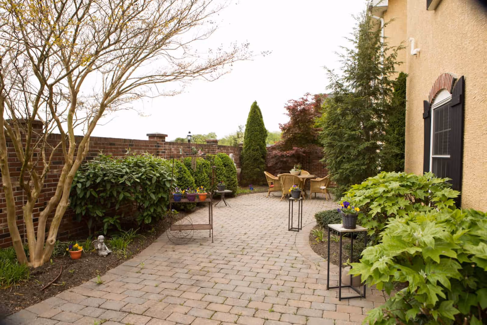 A paved outdoor patio area with a curved walkway surrounded by various green plants and bushes. There is a brick wall on the left side and a beige building wall with a window on the right. Several small tables with potted flowers are placed along the walkway, and a round table with chairs is visible in the background.