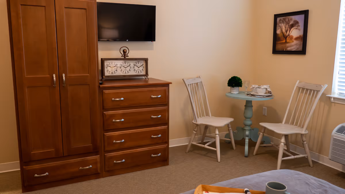 A cozy room with a wooden wardrobe and chest of drawers topped with a decorative clock and a wall-mounted flat screen TV. There is a small round table painted light blue with two white wooden chairs beside it. A framed landscape painting hangs on the wall above the table, and a window with blinds is partially visible. The corner of a bed with a gray blanket and a green cup on it is in the foreground.