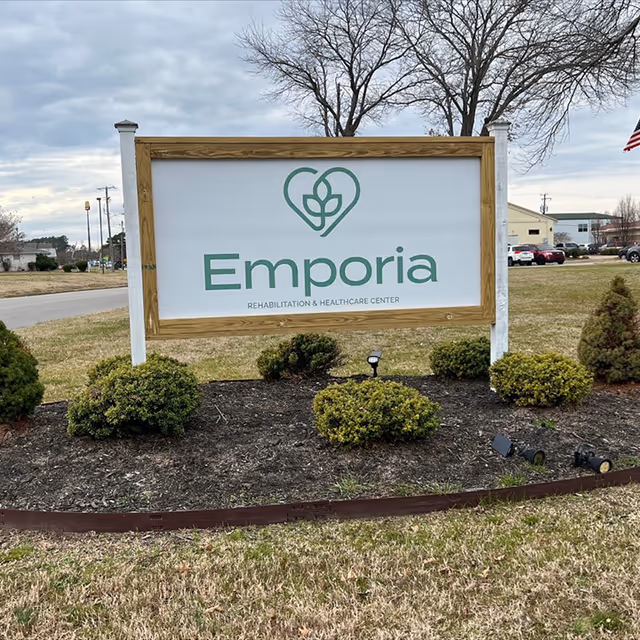 Outdoor sign for Emporia Rehabilitation & Healthcare Center with a heart and leaf logo, surrounded by small bushes and grass, with a cloudy sky and some buildings and cars in the background.