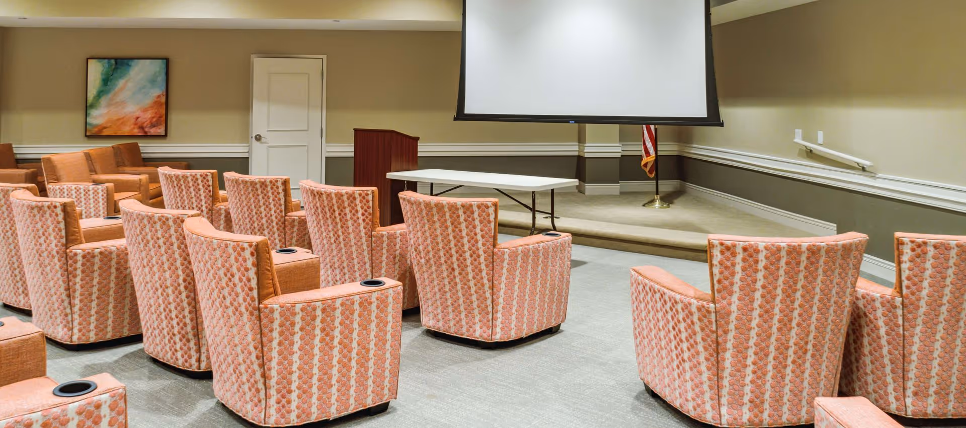 A small auditorium or meeting room with rows of patterned orange armchairs facing a stage area with a white projection screen, a podium, and a folding table. There is an American flag in the corner and a colorful abstract painting on the wall.