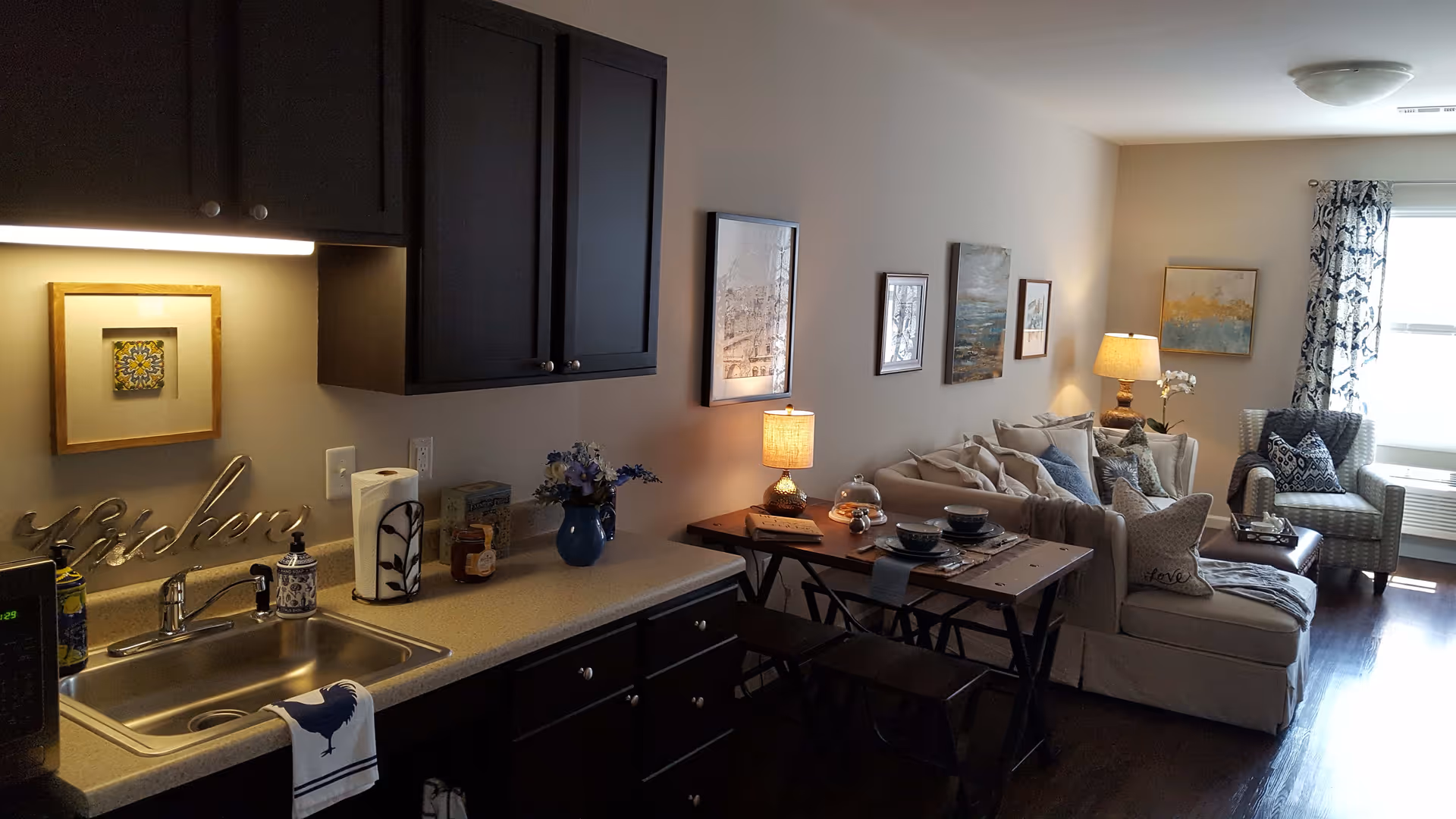 Interior view of a senior living facility unit at Vantage Pointe Village showing a small kitchen area with a sink, countertop, dark cabinets, and decorative items. Adjacent to the kitchen is a cozy living room with a beige sectional sofa, a wooden dining table with place settings, a cushioned armchair near a window with patterned curtains, and several framed artworks on the walls. The room is softly lit by table lamps.