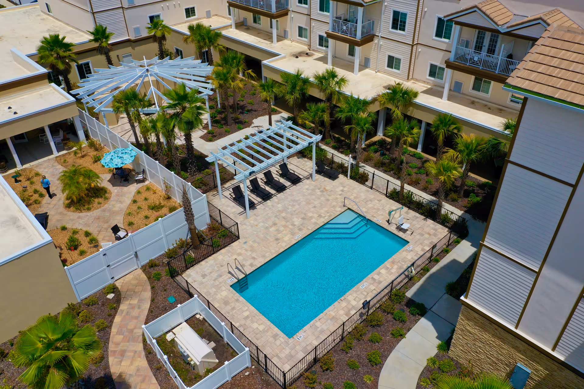 Aerial view of an outdoor swimming pool area at Seagrass Village of Fleming Island, surrounded by a paved deck with lounge chairs under a pergola, palm trees, landscaped garden beds, and adjacent buildings with balconies.