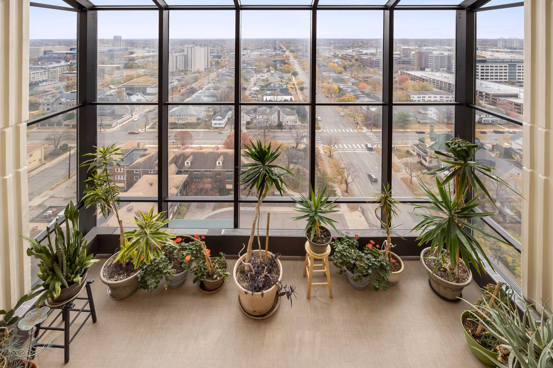 Indoor garden area with various potted plants arranged near a large floor-to-ceiling window offering a panoramic view of a cityscape with buildings, streets, and trees.