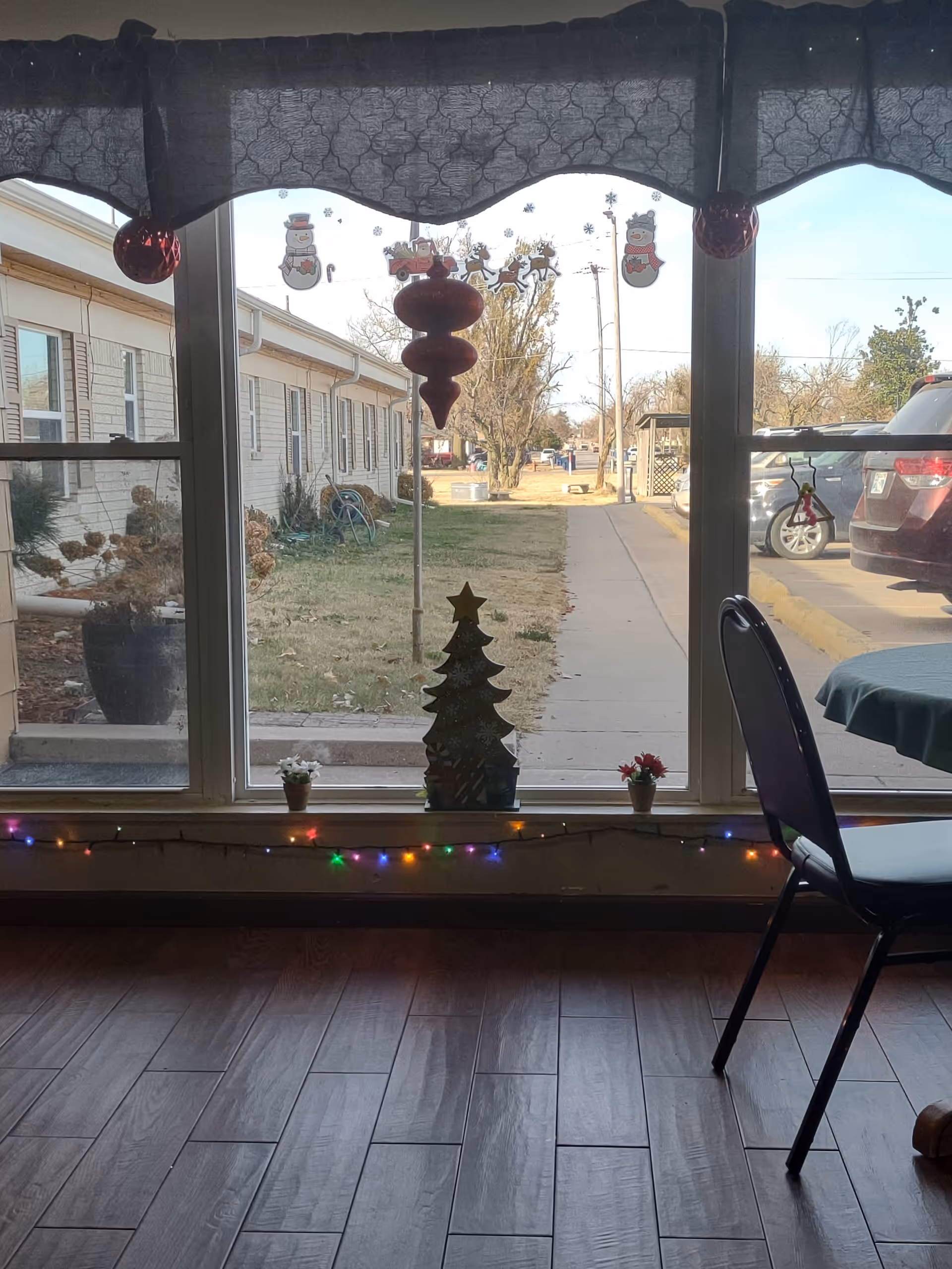 Interior view of a common area with a decorated windowsill and Christmas ornaments looking out onto a sidewalk and parked cars, with a chair and table at the right.