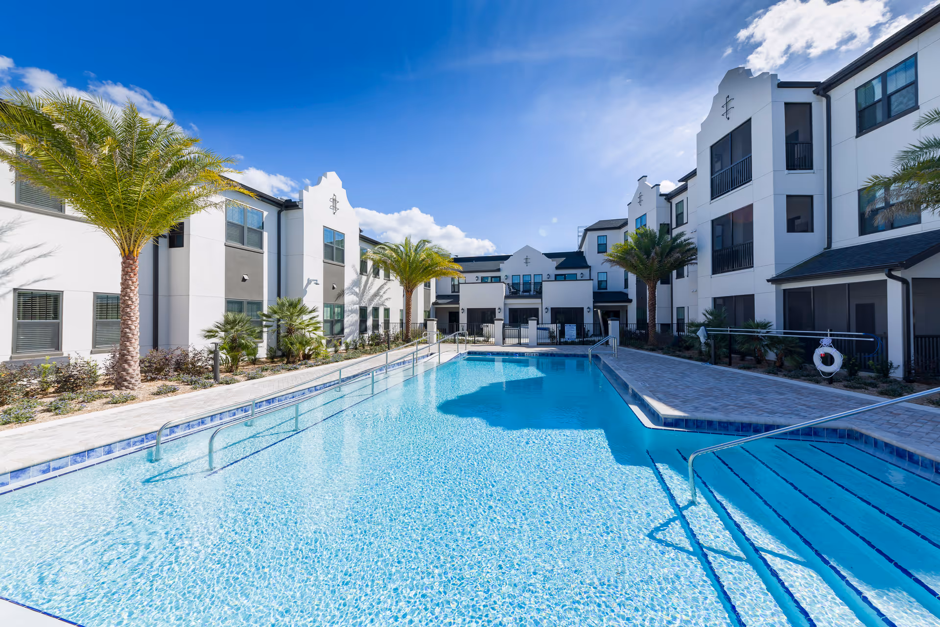 Outdoor swimming pool with clear blue water surrounded by a paved deck and palm trees, adjacent to a modern three-story white building under a bright blue sky with some clouds.
