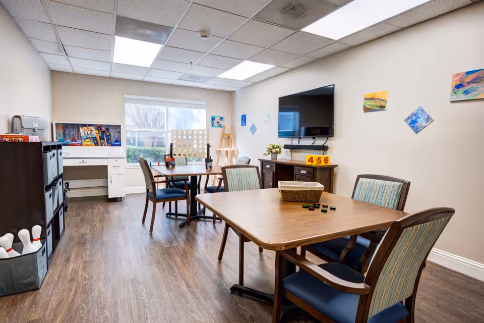 A bright activity room with wooden floors featuring two tables surrounded by chairs. On one table, there are game pieces and a basket, while the other table has a large Connect Four game. A flat-screen TV is mounted on the wall above a wooden cabinet with a flower vase and numbered blocks. The walls are decorated with colorful artwork, and a window lets in natural light. Shelving units and a desk are visible along the left wall.