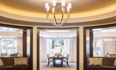 A bright and elegant interior space featuring a central dining area with a rectangular table and chairs, flanked by two seating areas with cushioned benches. The room has large windows with sheer curtains allowing natural light to fill the space, and modern chandeliers hanging from the ceiling.