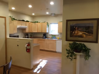 A bright kitchen area with light wood cabinets, a white dishwasher, and a coffee maker on the counter. There is a small window above the sink letting in natural light. The kitchen is open to an adjacent room with a partial wall and a fire extinguisher mounted on it. A framed painting and a potted plant on a pedestal are visible on the right side of the image.