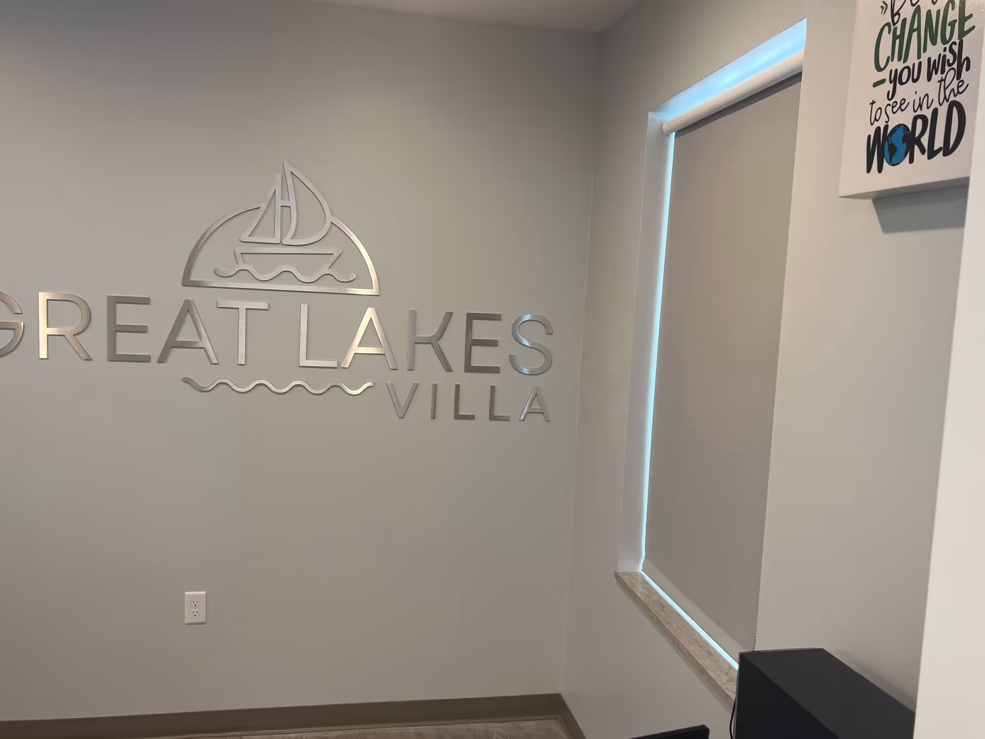 Interior wall with metallic letters spelling 'GREAT LAKES VILLA' and a sailboat logo above the text. To the right, there is a window with a closed blind and a partial view of a framed quote on the wall.