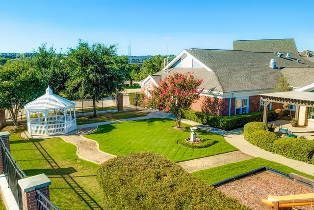 A well-maintained outdoor garden area at The Auberge at Valley Ranch featuring a white gazebo, green lawns, paved walkways, trimmed bushes, and a brick building with a covered patio. Trees and a fence are visible in the background under a clear blue sky.