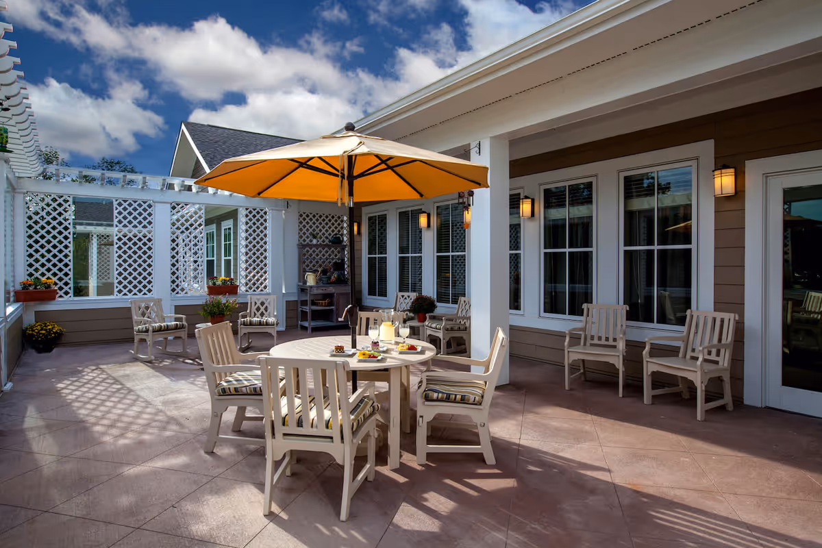 Outdoor patio with a round dining table and chairs under a yellow umbrella beside building windows and lattice privacy panels.