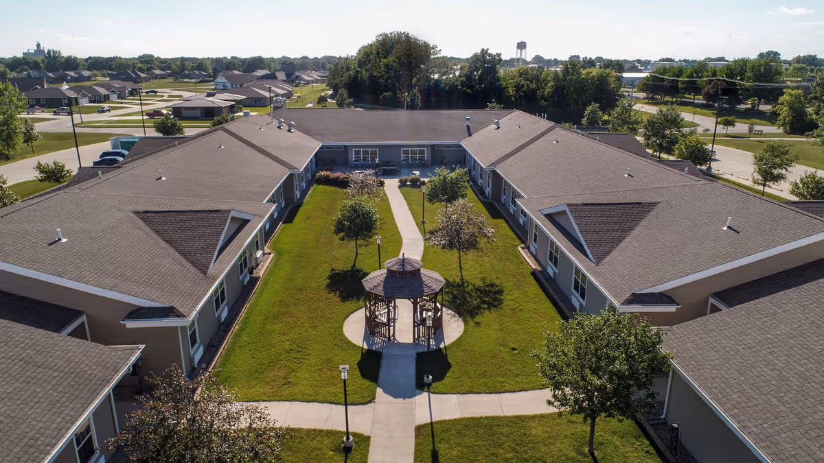 Aerial view of Homestead Assisted Living & Memory Care of Algona showing a U-shaped building surrounding a green courtyard with a gazebo in the center. The courtyard has a paved walkway, small trees, and lamp posts. Residential houses and trees are visible in the background under a clear sky.