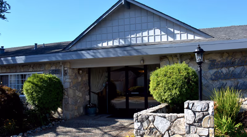 Front exterior view of Brookside Skilled Nursing Hospital showing a stone facade building with a peaked roof, glass double doors entrance, two neatly trimmed round bushes on either side of the entrance, a stone wall with a lamp post, and some greenery around the pathway leading to the door.