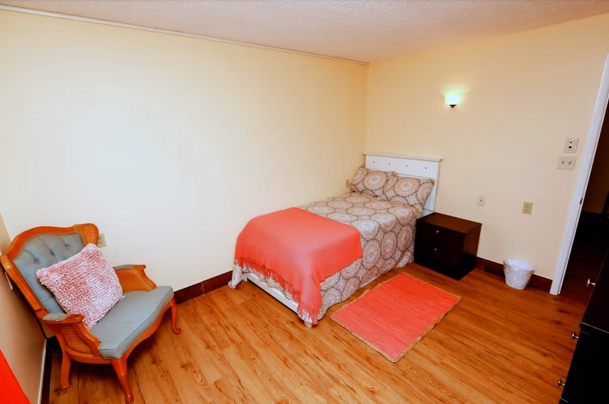 A small, cozy bedroom with a single bed covered in patterned bedding and a coral throw blanket. Next to the bed is a dark wooden nightstand and a small coral rug on the wooden floor. There is a vintage-style armchair with a pink cushion in the corner. The walls are painted light beige, and a wall-mounted light fixture is above the bed.