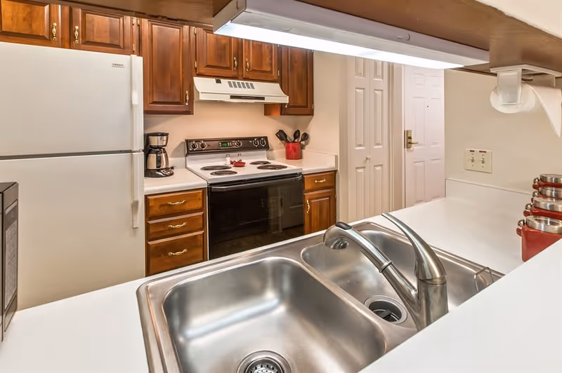 A kitchen with a double stainless steel sink in the foreground, white countertops, wooden cabinets, a white refrigerator, an electric stove with an oven, a coffee maker, and a set of red canisters on the counter.