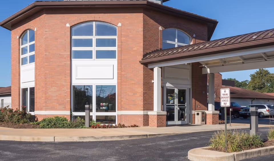 Exterior view of a brick building with large windows and a covered entrance. There is a driveway in front with a sign indicating parking for emergency vehicles only. Some landscaping with bushes and flowers is visible near the building.