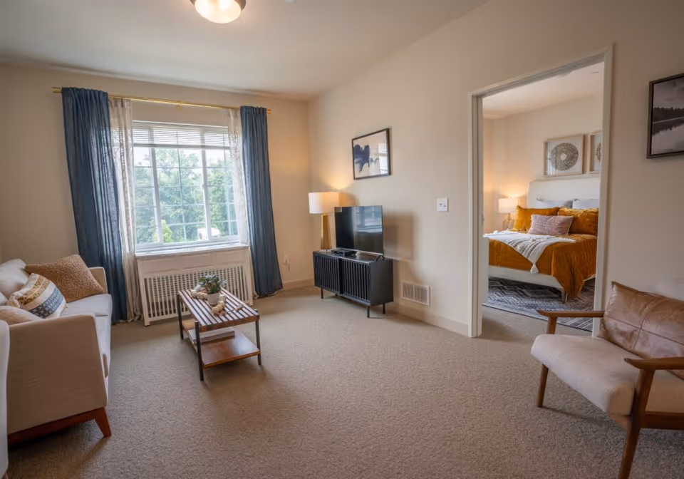 A cozy living room with a beige sofa, a wooden coffee table with decorative items, a TV on a black stand, and a floor lamp. The room has a large window with blue and white curtains letting in natural light. Through an open doorway, a bedroom is visible with a bed covered in mustard yellow bedding and decorative pillows, bedside tables with lamps, and wall art.