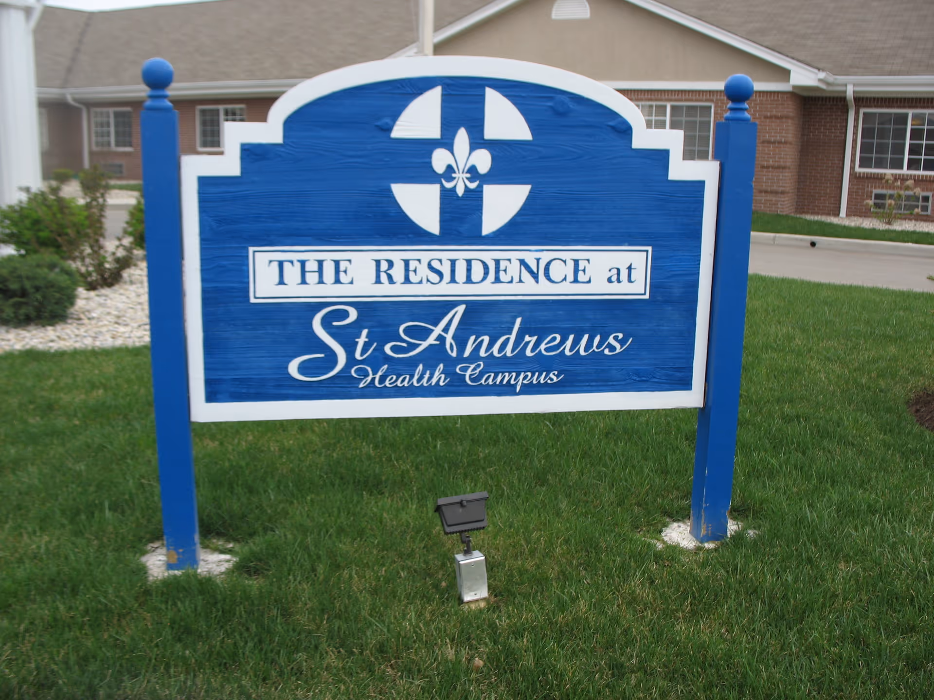 Blue and white wooden sign on green grass in front of a brick building, displaying the text 'THE RESIDENCE at St Andrews Health Campus' with a decorative emblem above the text.
