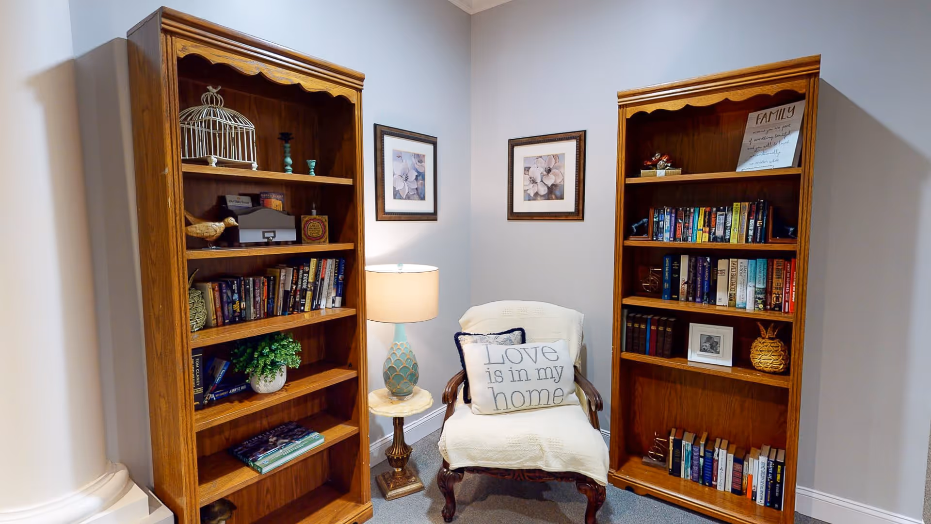 A cozy reading nook featuring two wooden bookshelves filled with books and decorative items, a cushioned armchair with a pillow that reads 'Love is in my home', a small round side table with a lamp, and two framed floral pictures on the wall.