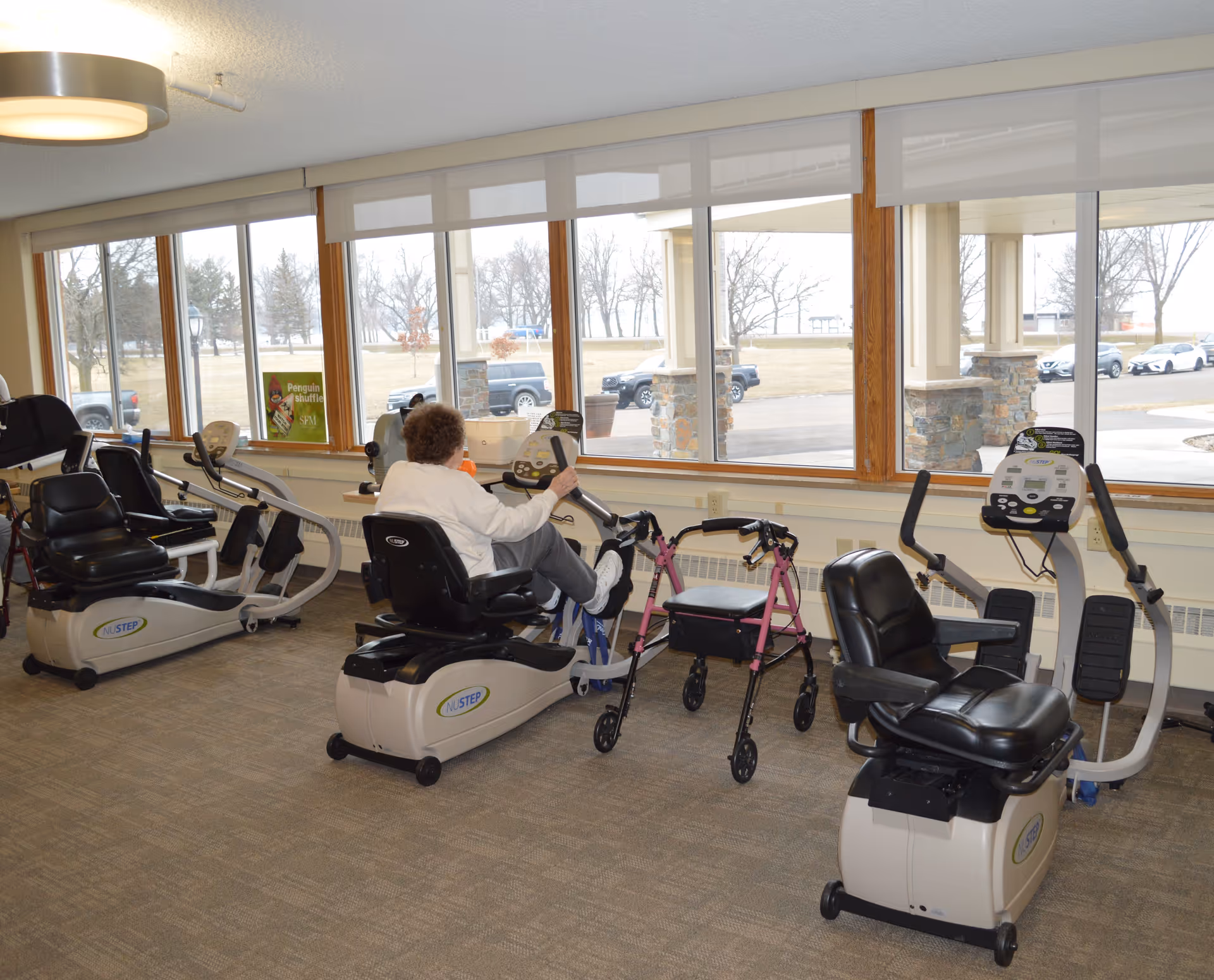 An elderly person using a recumbent exercise bike in a fitness room with large windows showing an outdoor view. Several other exercise machines and a pink walker are also visible in the room.
