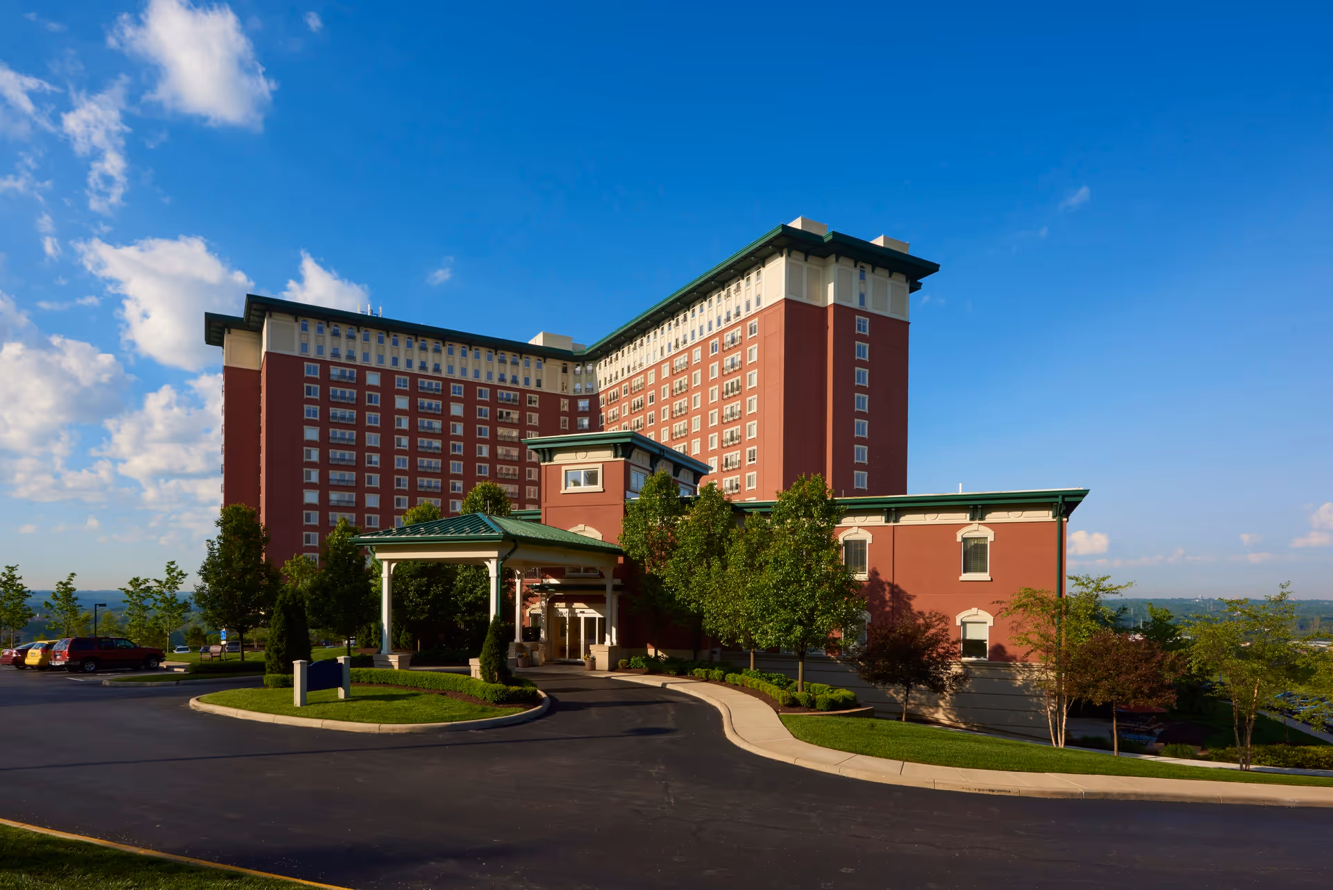 Exterior view of a large multi-story senior living facility building with red brick walls and green roof accents under a clear blue sky. The building is surrounded by trees and a paved driveway with parked cars.