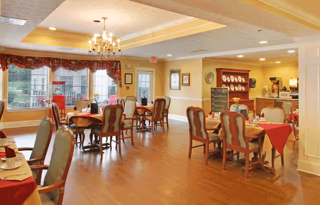A warm and inviting dining room with several wooden tables and chairs arranged neatly. The tables are set with red tablecloths, napkins, and white dishware. Large windows with floral valances allow natural light to fill the room. A chandelier hangs from the ceiling, and a wooden hutch with plates and a chalkboard menu is visible in the background near a coffee station.