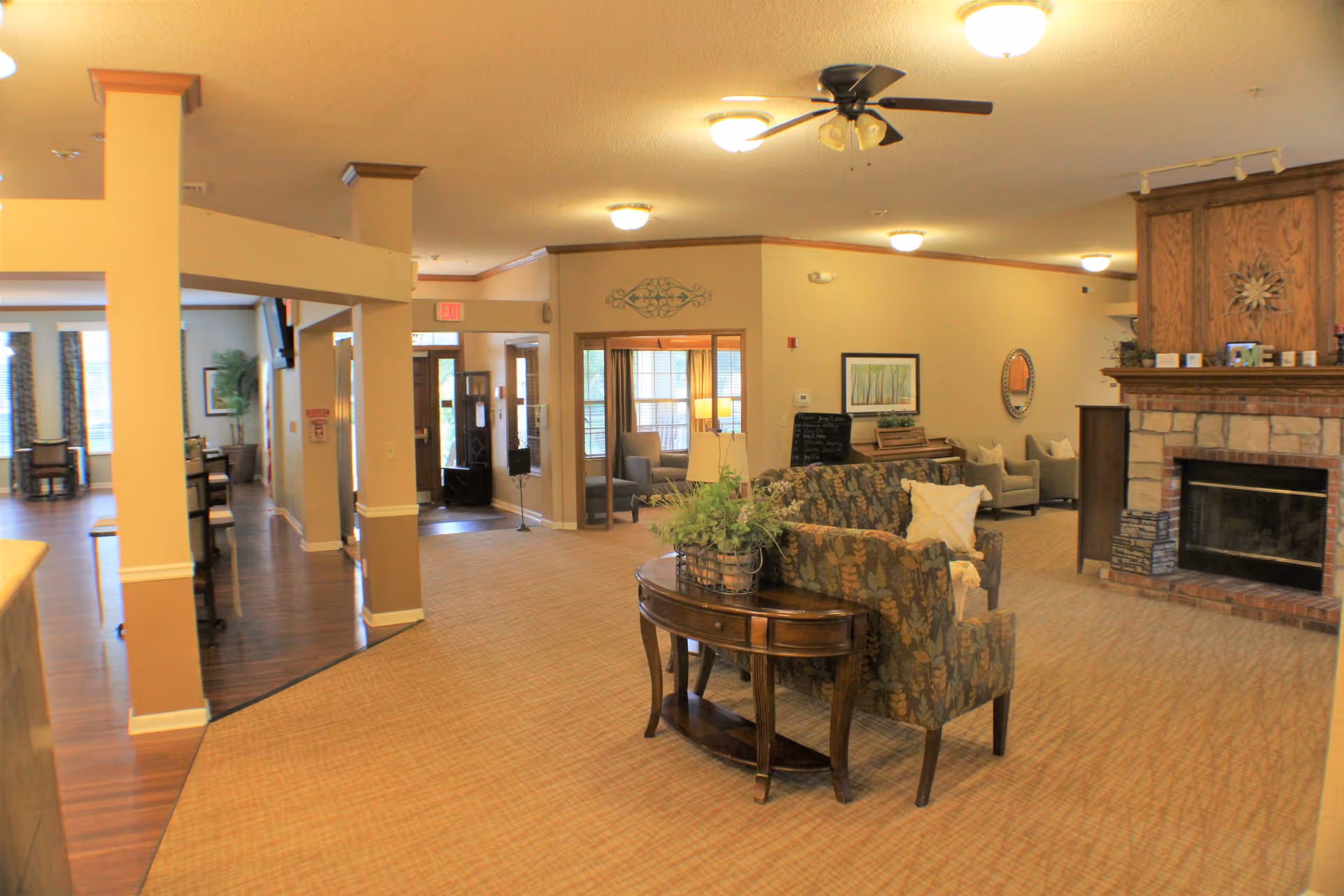 A spacious and warmly lit common area in a senior living facility featuring a patterned sofa with a white pillow, a wooden console table with a plant, a stone fireplace with a wooden mantle decorated with the word 'LOVE', and several armchairs. The room has beige walls, carpeted floors, ceiling lights, and a ceiling fan. In the background, there is a hallway leading to an entrance door and another seating area with large windows.