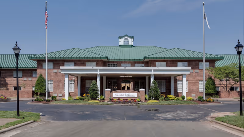 Front exterior view of Bartlett Reserve, a brick building with a green metal roof, two flagpoles with flags, and a covered entrance with landscaping and a sign displaying the facility name.