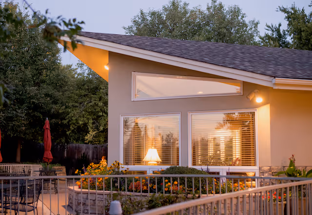 Exterior view of a single-story building at dusk with large windows showing a lit lamp inside. The building has a sloped roof and is surrounded by a garden with colorful flowers and greenery. There is a metal railing and outdoor seating area with tables and chairs visible in the foreground.