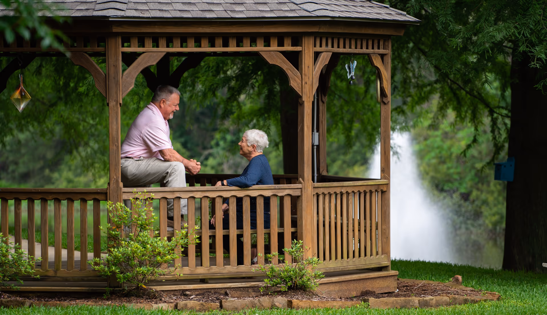 An elderly man and woman sitting and talking inside a wooden gazebo surrounded by greenery with a water fountain visible in the background.