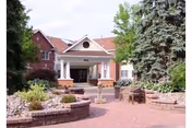 Front entrance of a brick senior living building with a covered portico, columns, brick-paved driveway and landscaped planters.