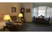 A cozy living room area with a recliner chair, two side tables each with a lamp, a grandfather clock, framed pictures on the wall, and a window with blinds and a valance. There is also a small table with two chairs near the window.