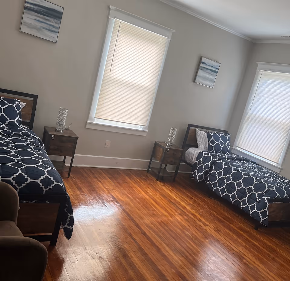 Bright bedroom with two twin beds in navy patterned bedding, wooden floors, two nightstands with lamps, and windows with blinds.