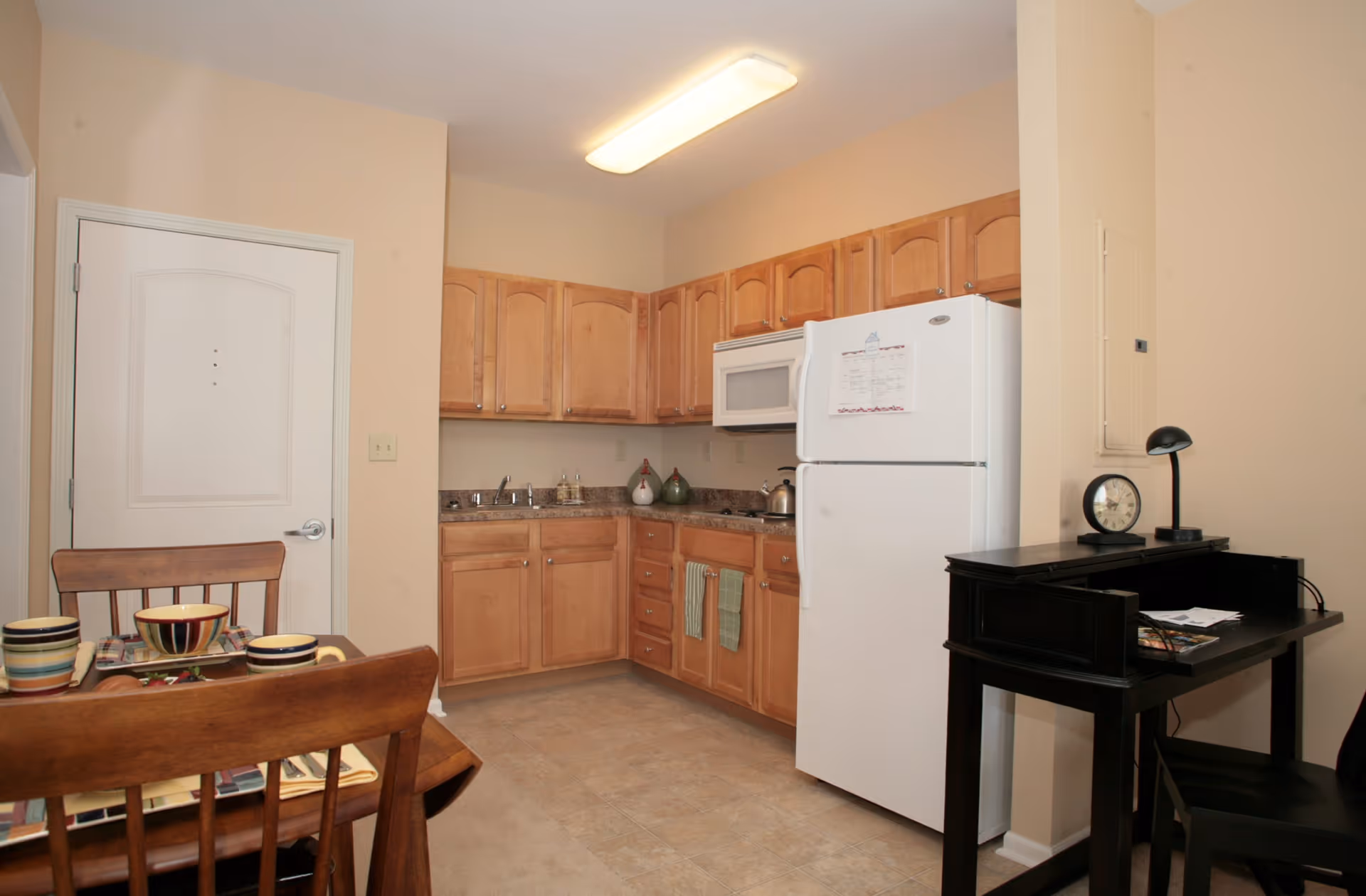 A small kitchen area with wooden cabinets, a white refrigerator, a microwave, and a countertop with a sink. To the left, there is a wooden dining table set with colorful bowls and plates. On the right side, there is a black desk with a clock, a lamp, and some papers on it. The walls are beige and the floor is tiled.