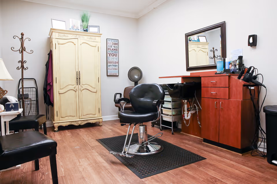 Interior of a small salon room with a black salon chair on a black mat in the center, a wooden cabinet with drawers and a sink on the right, a vintage cream-colored armoire against the back wall, a coat rack with hanging clothes, a hair dryer mounted on the wall, and a large mirror above the cabinet. The room has wooden flooring and light-colored walls.