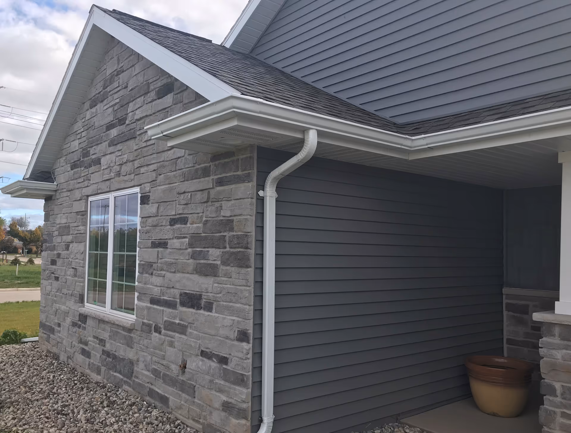 Stone and gray vinyl sided exterior corner of a house with a window, gutters, and a covered porch with a planter.