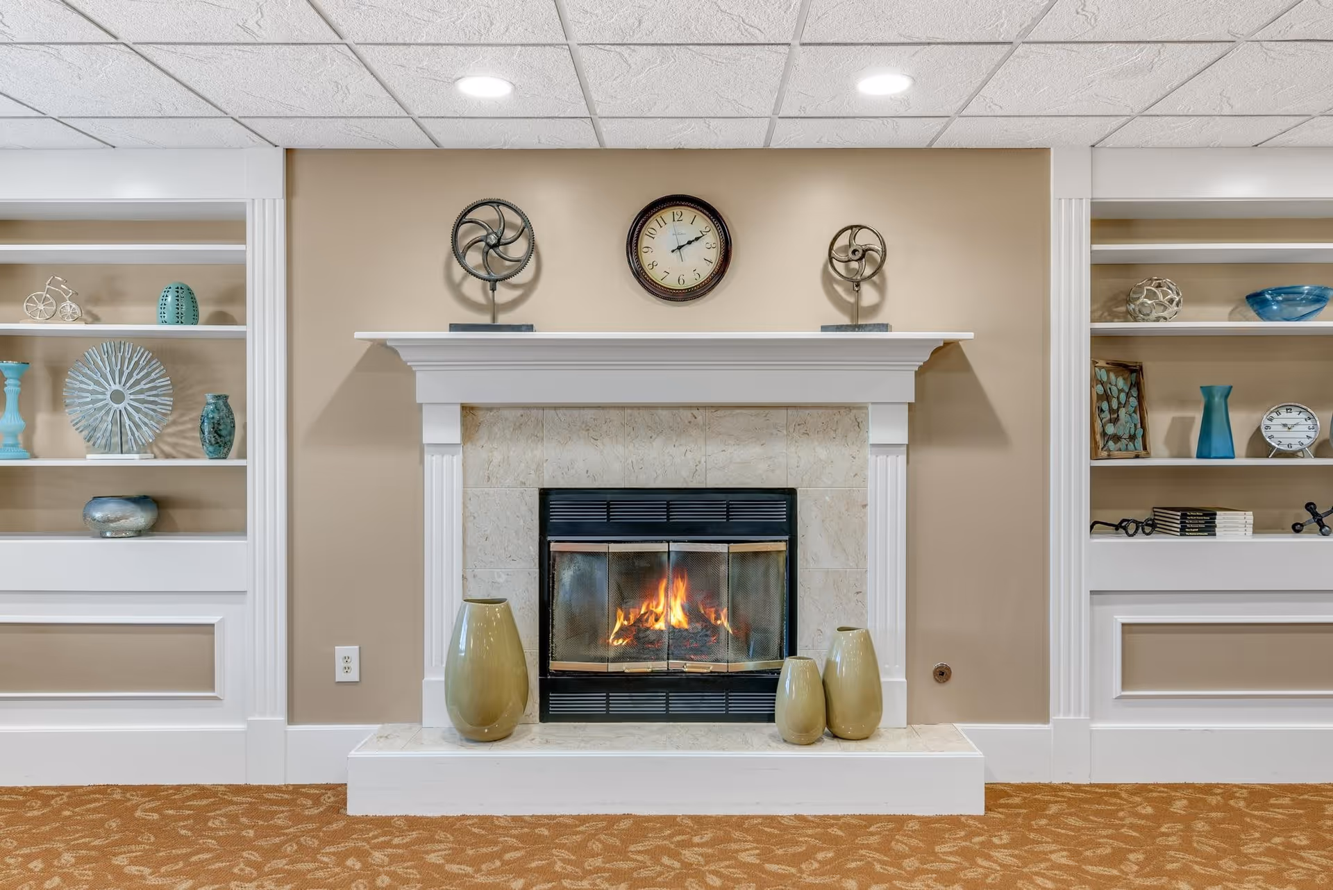 A cozy indoor fireplace with a fire burning, surrounded by a white mantel and beige tiled surround. Above the mantel is a round clock showing the time as 11:05, flanked by two decorative metal fan sculptures. On the hearth are three ceramic vases of varying sizes. Built-in white shelves on either side of the fireplace display various decorative items including vases, books, and small sculptures. The walls are painted beige and the floor is covered with a patterned carpet.