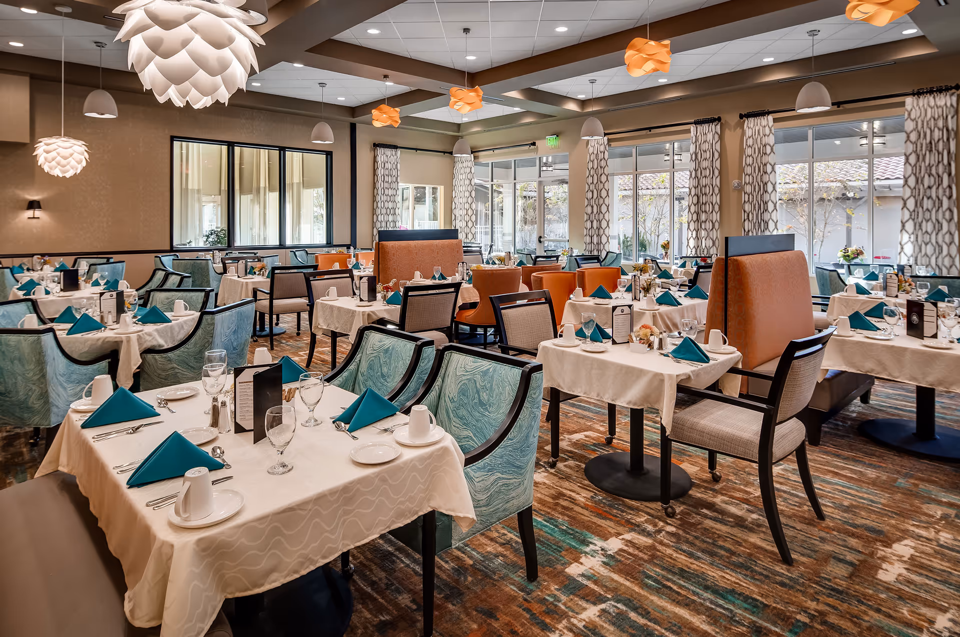 Spacious, well-lit dining room with multiple tables set with white linens and teal napkins, patterned chairs, and large windows.