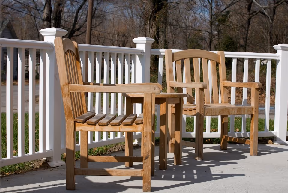 Two wooden chairs and a small wooden table on a sunlit porch with a white railing and trees beyond.