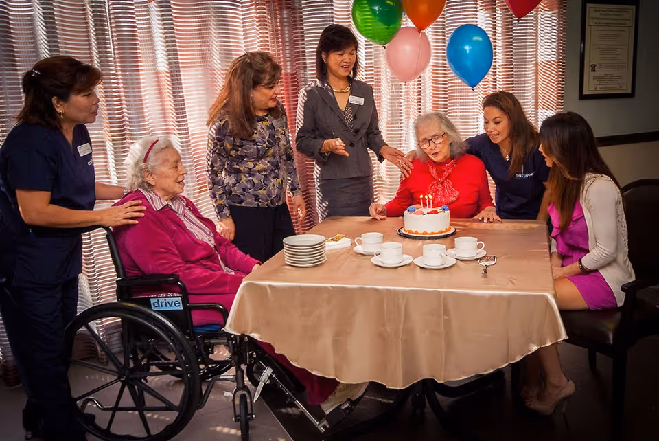 A group of six women gathered around a table celebrating a birthday. One elderly woman in a red sweater is seated at the table with a birthday cake with lit candles in front of her. Another elderly woman in a wheelchair wearing a magenta jacket is also present. The other four women, likely staff or family, are standing or sitting around the table smiling. Colorful balloons are tied behind the elderly woman with the cake. The setting appears to be a cozy indoor room with curtains in the background.