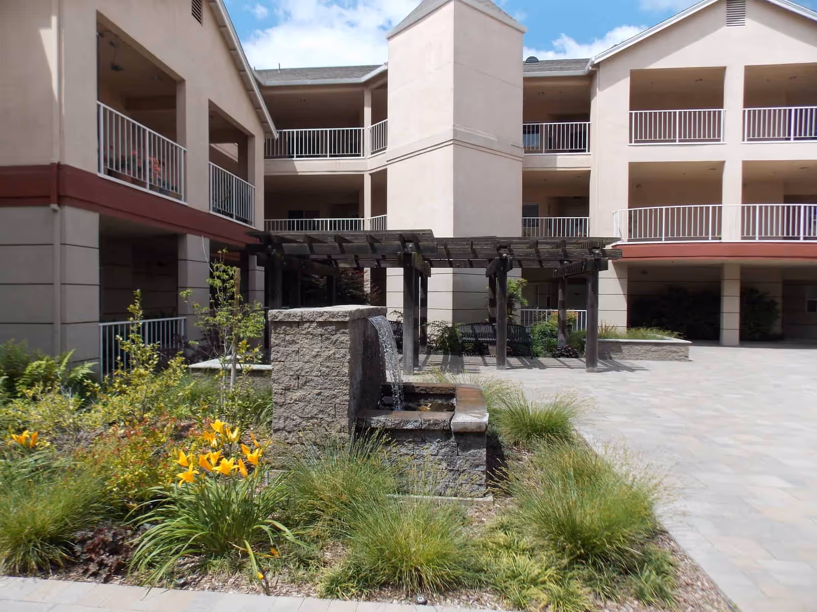 Outdoor courtyard area of a senior living facility with a small stone water fountain, yellow flowers, green plants, and a wooden pergola with benches underneath. The building surrounding the courtyard has beige walls and multiple balconies with white railings.
